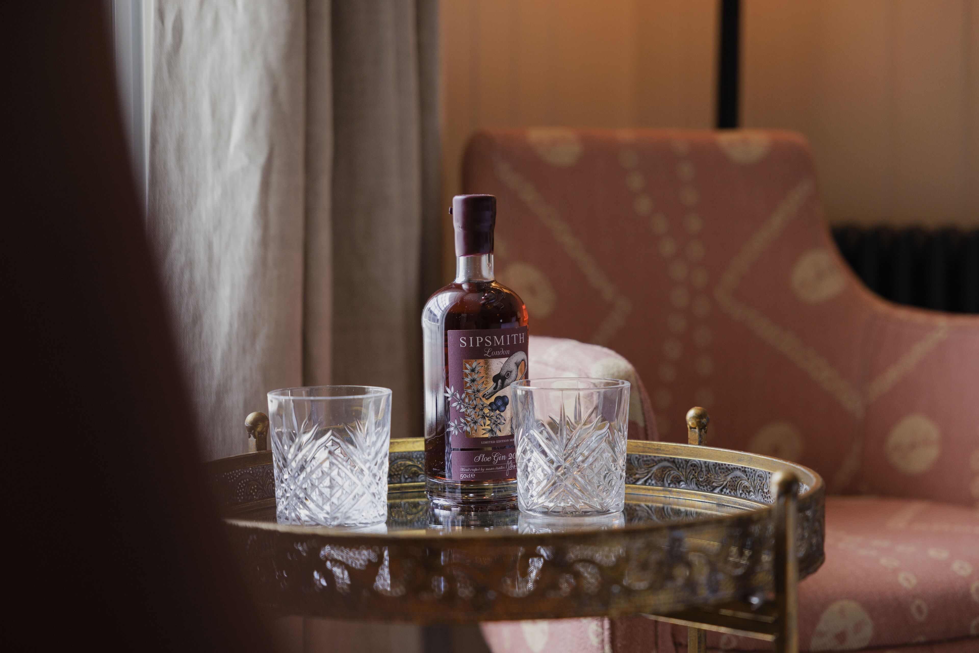 A bottle of Sipsmith gin, two crystal glasses, and a patterned armchair on a table.