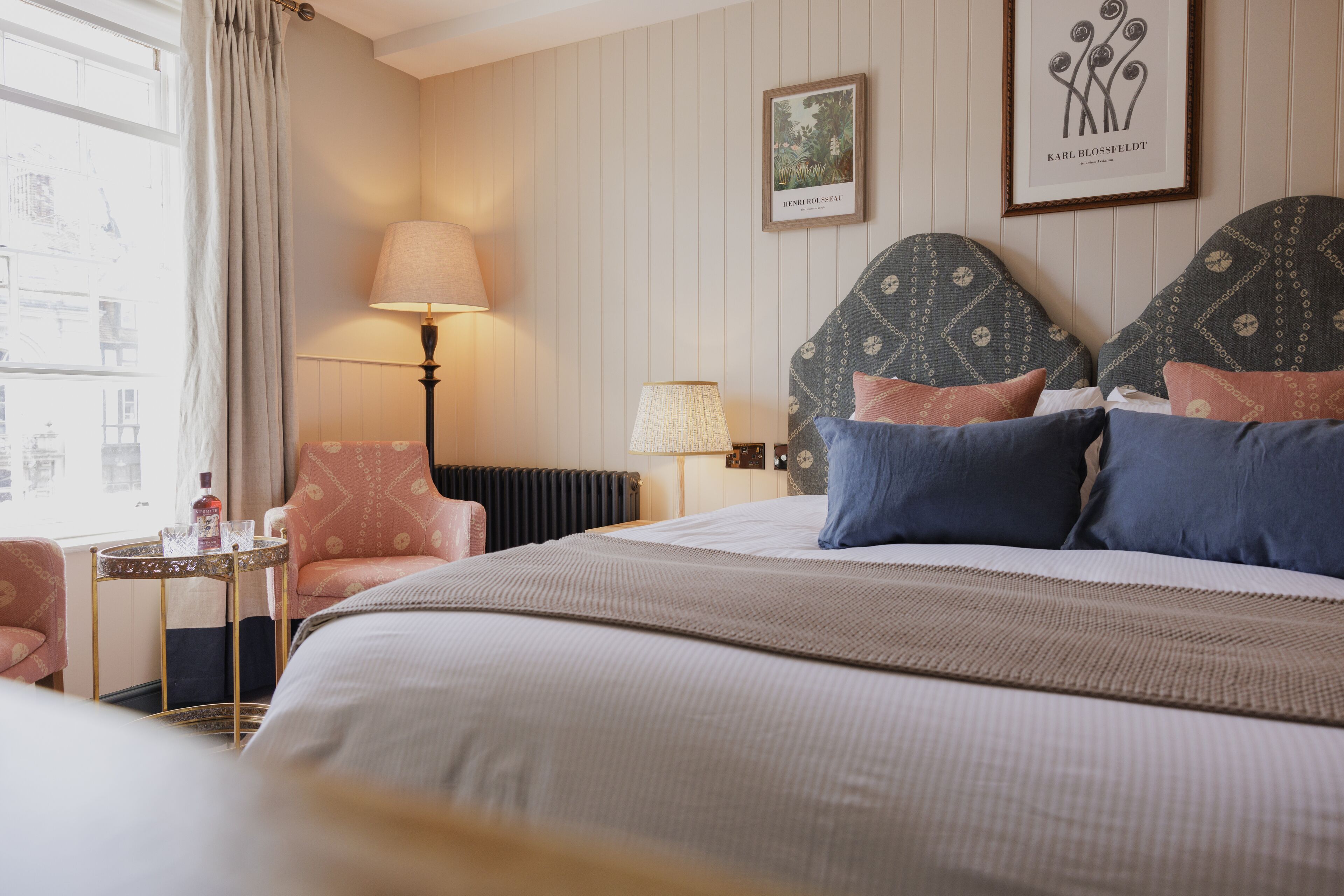 Habitación de hotel con una cama, dos sillas, una mesa, una lámpara y dos cuadros en la pared.