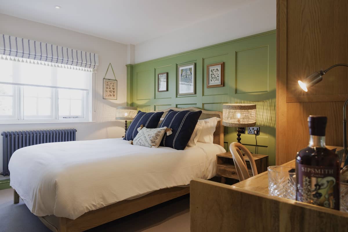 A neatly made bed with white bedding and blue pillows, a bedside table with a lamp, and a bottle of Sipsmith on the nightstand.