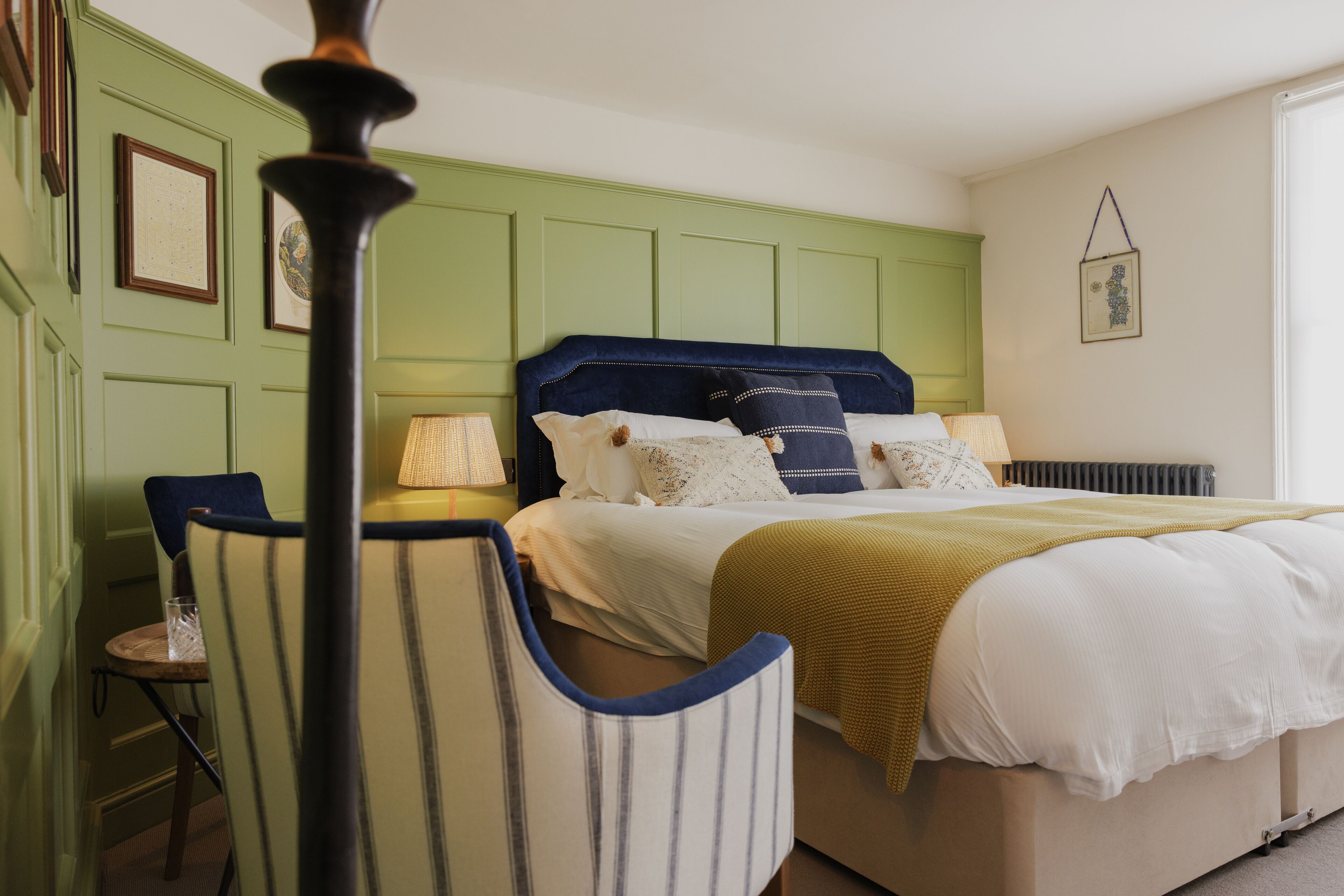 A bedroom with a green paneled wall, a bed with white bedding and a blue headboard, a yellow throw blanket, and a chair with striped upholstery.