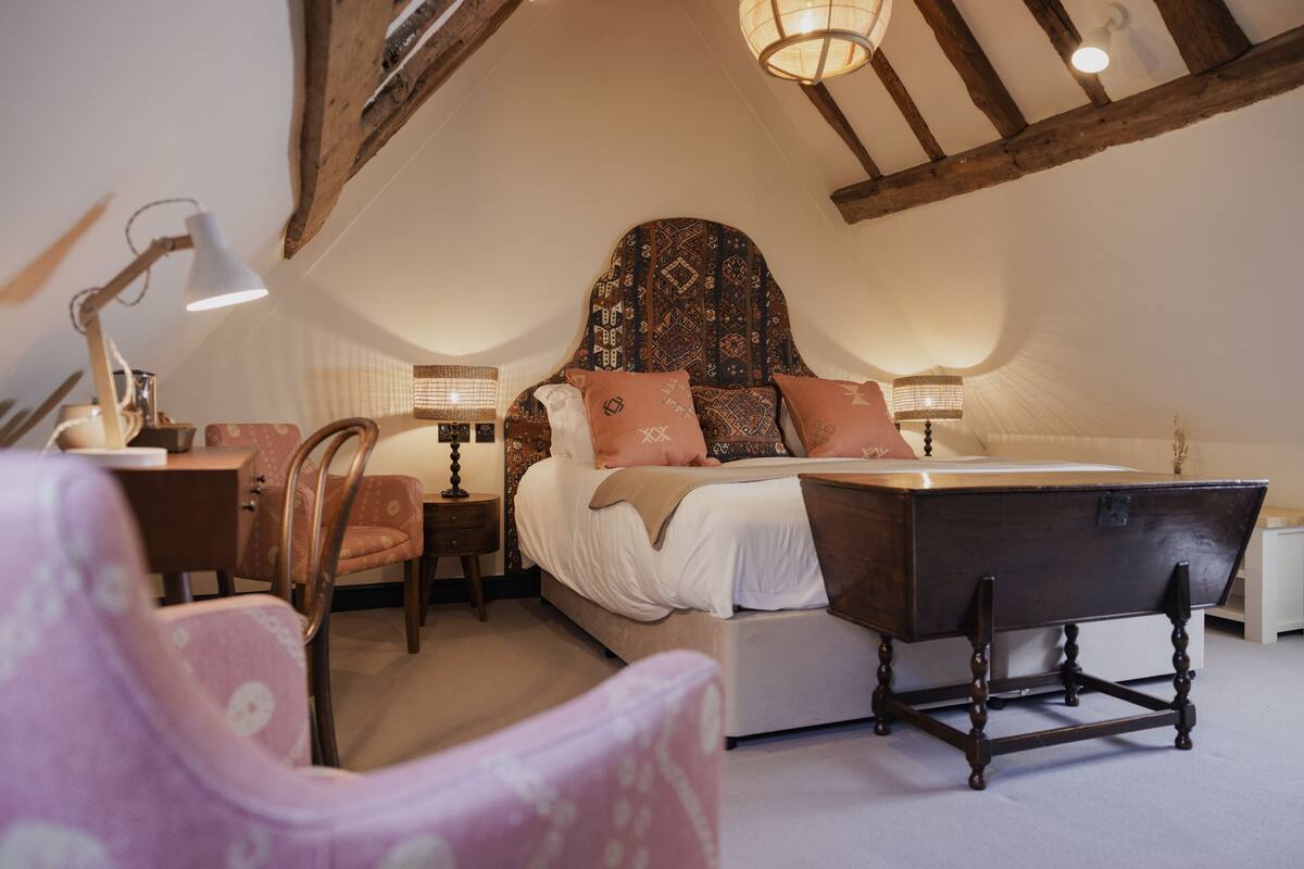 A bedroom with a large bed, a wooden bench, a chair, and a desk with a lamp.