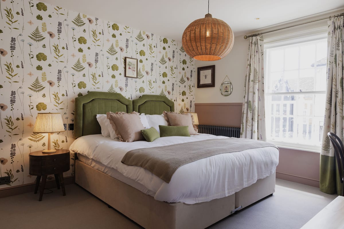 A bedroom with a large bed, a nightstand with a lamp, floral wallpaper, and a window with curtains.