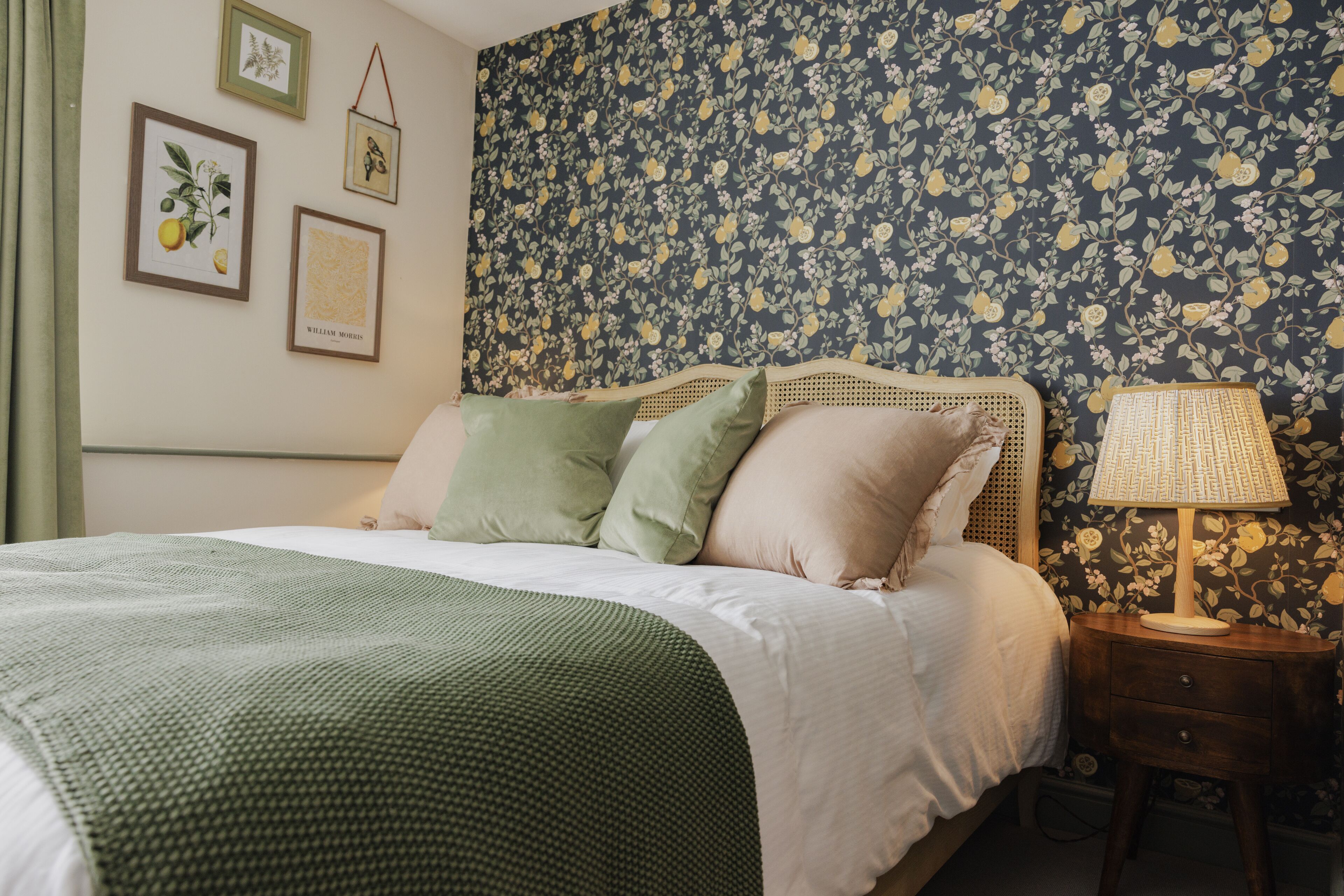 Un letto rifatto con una coperta verde, un comodino in legno con una lampada e carta da parati a motivi floreali.