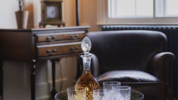 A room with a dark wooden table, a glass decanter, and crystal glasses on a tray. There is a lamp, a clock, and a plant in the background.