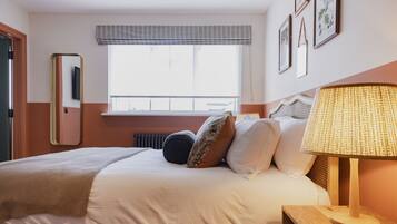 A neatly made bed with a white comforter and pillows, a bedside lamp, and a window with a view of the outside.