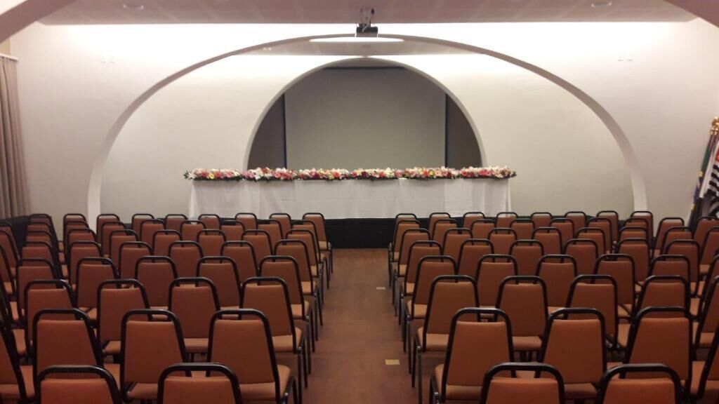Un salón de conferencias con filas de sillas mirando hacia un escenario con un arreglo floral.