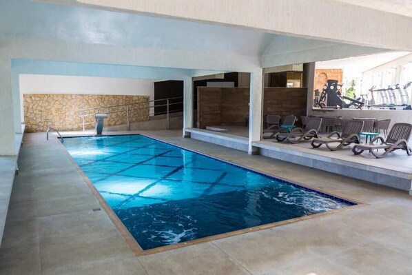 An indoor swimming pool with a stone wall backdrop, a diving board, and a seating area with chairs and tables.
