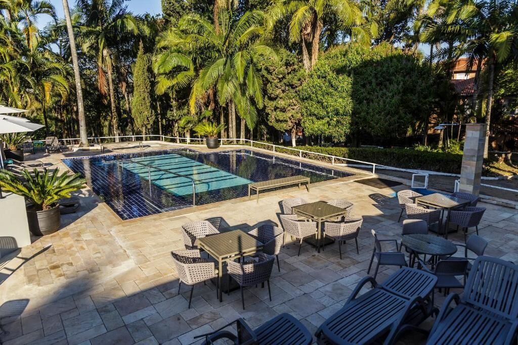 Uma piscina retangular com borda azulejada, rodeada por espreguiçadeiras e mesas num terraço de pedra, com palmeiras e vegetação densa ao fundo.
