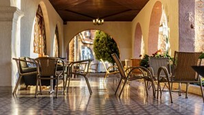 A covered outdoor seating area with wicker chairs and a tiled floor, leading to an archway with a view of greenery and buildings.