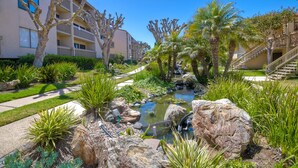 A landscaped garden with a small water feature, palm trees, and a building with balconies. - Ocean Dreams – Cozy Family Condo Near Beach, Pool & Tennis in Solana Beach (Solana Beach)