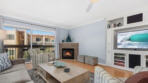 A living room with a fireplace, a television, and a balcony with outdoor furniture. - Ocean Dreams – Cozy Family Condo Near Beach, Pool & Tennis in Solana Beach (Solana Beach)
