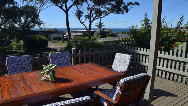 Une terrasse en bois avec une table et des chaises, une clôture blanche en bois et une vue sur l’océan.