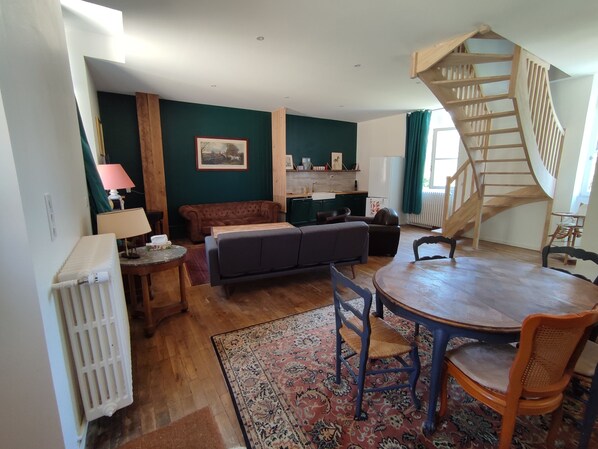 Een gezellige woonkamer met een houten trap, een bank, een eettafel met stoelen, en een keuken in de achtergrond.