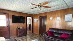 A living room with a brown leather sofa, a television mounted on the wall, a ceiling fan, and a wooden paneled wall.