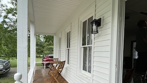 A covered porch with wooden chairs and a table, a red car parked outside, and a house with white siding and windows.