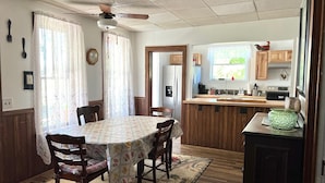 A dining area with a round table, chairs, a ceiling fan, and a kitchen with wooden cabinets and a window.