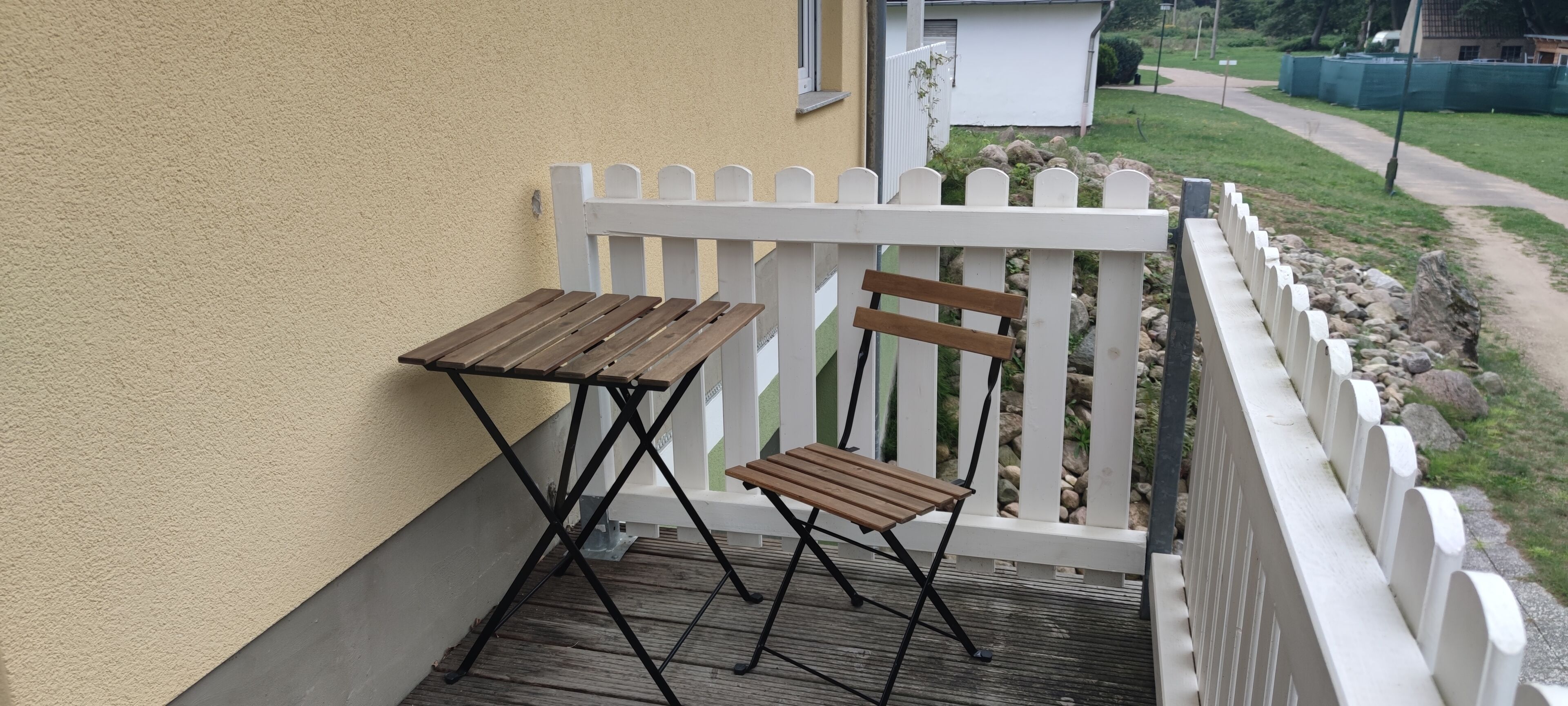 En balkong med ett träbord och en stol, ett vitt staket och utsikt över en trädgård och en gångstig.