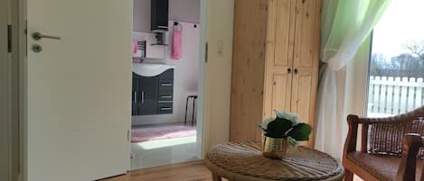 A room with a wooden floor, a wicker chair, a small table with a plant, and a wardrobe.