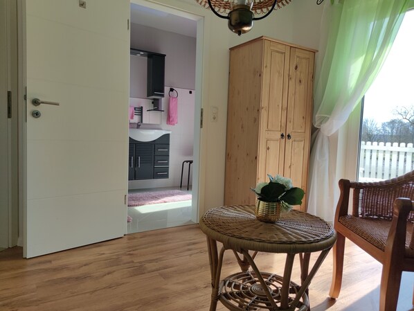 Una habitación con piso de madera, una silla de mimbre, una pequeña mesa con una planta y un armario.