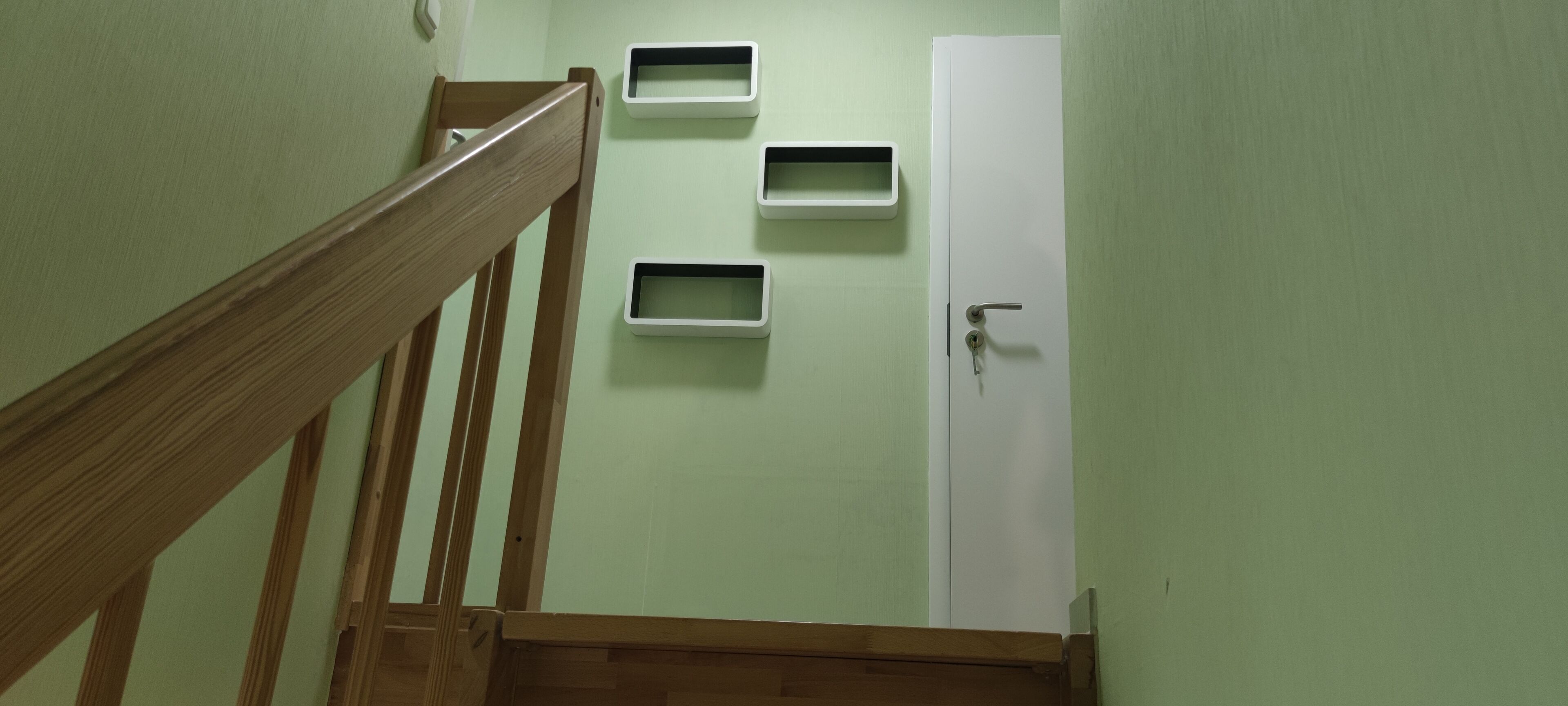 Eine Treppe mit hölzernen Geländern führt zu einer Tür mit Schlüsselloch. An der Wand befinden sich drei weiße, rechteckige Stauraumfächer.