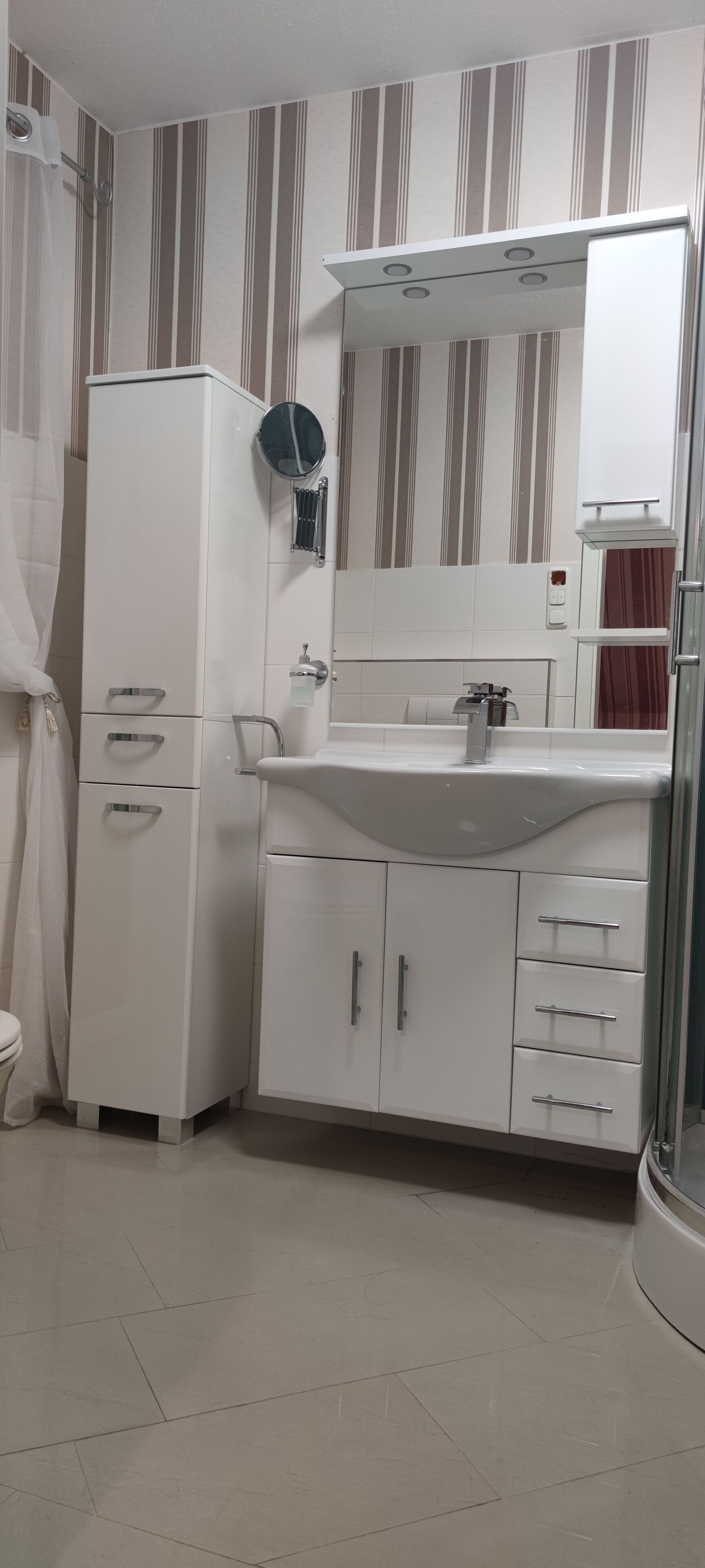 Ein Badezimmer mit weißem Waschbecken, Spiegel und Schrank. Eine Duschvorhang und eine Toilette sind sichtbar.