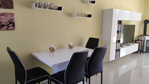A modern dining area with a white table and black chairs, white shelves with wine glasses, and a white cabinet with books.
