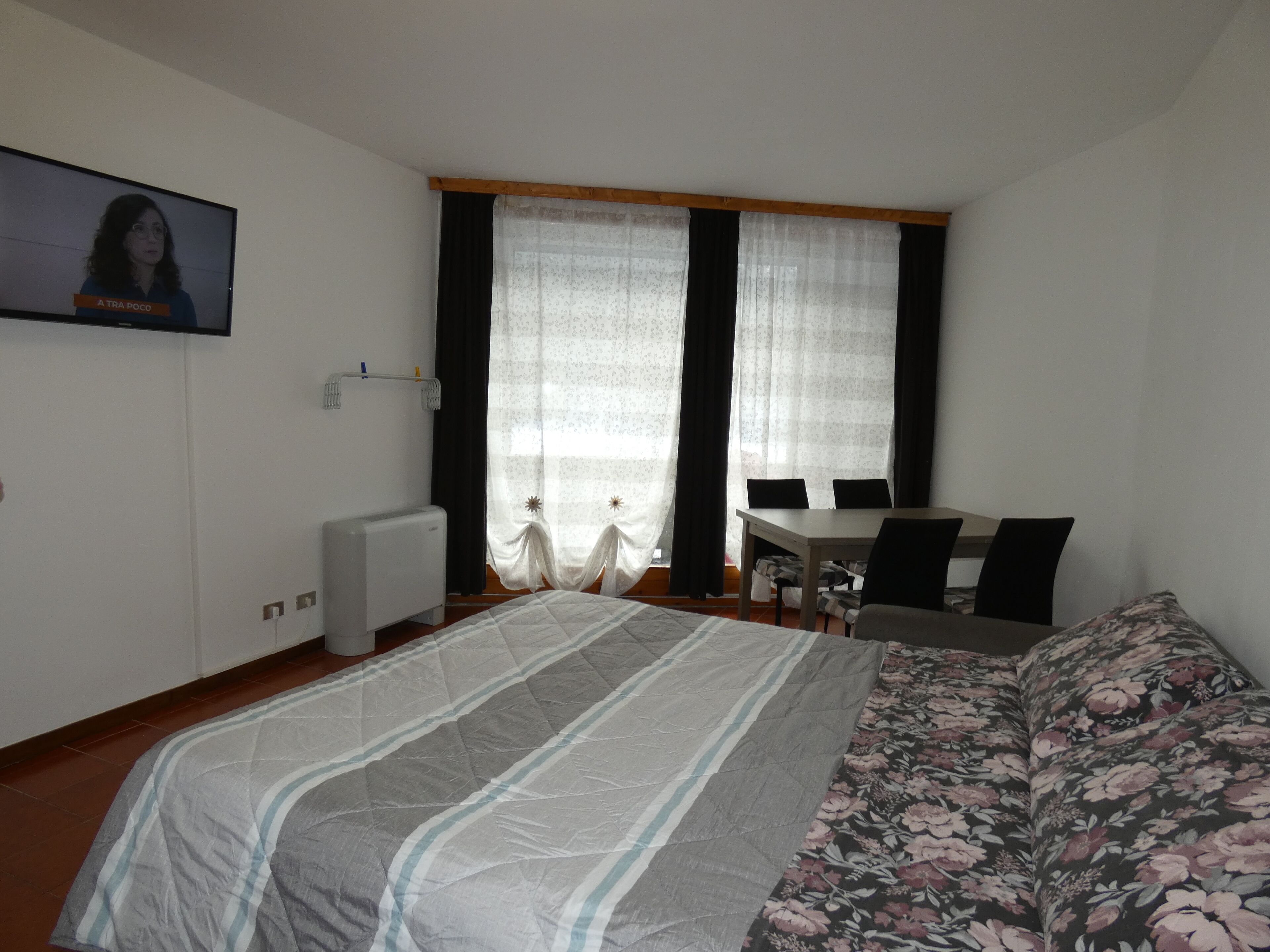 Une chambre à coucher avec un lit, un bureau et une chaise. Il y a une fenêtre avec des rideaux et un radiateur au mur.