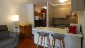 A compact living space with a kitchenette, a sofa, a dining area with stools, and a bedside table with a lamp.