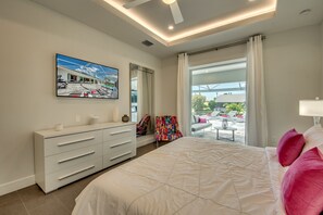 A modern bedroom with a large bed, a white dresser, a colorful chair, and a view of the outdoors through a large window. - Villa Seven Oceans - stylish new villa, extra large pool & spa, boat dock (Cape Coral)