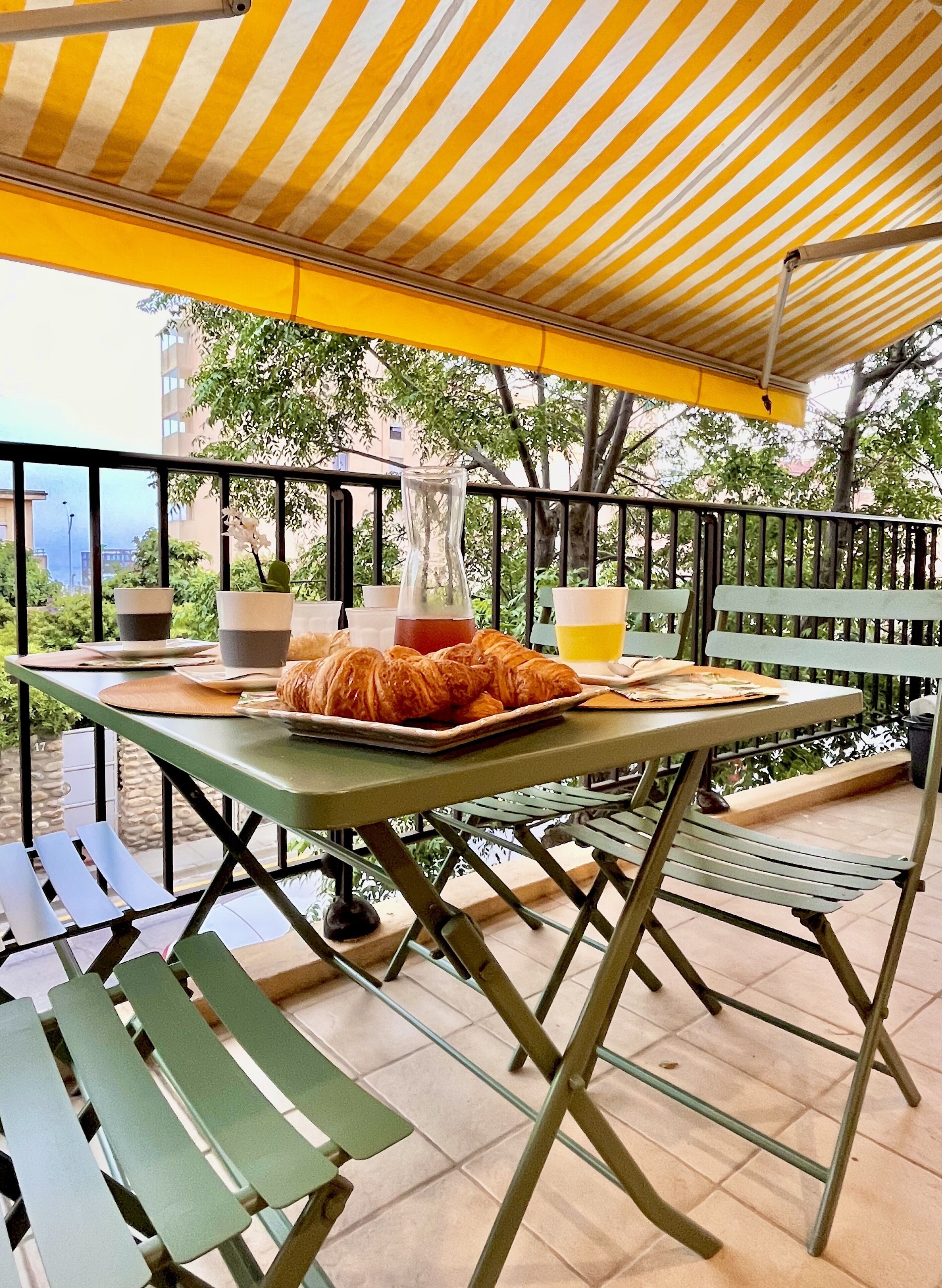 Ein Essbereich im Freien mit Tisch, gedeckt für das Frühstück, einschließlich Croissants und einem Saftkrug, unter einer Streifenmarkise.