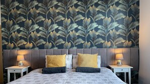 A neatly made bed with a floral bedspread, two matching pillows, and a headboard with a leaf-patterned wallpaper. There are two bedside tables with lamps on either side of the bed. - Between the beach and the shops (Portsmouth)