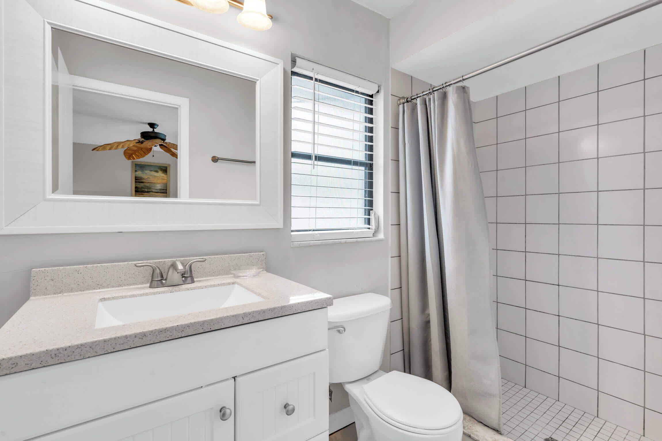 A bathroom with a white sink, a toilet, a shower with a curtain, and a window with blinds.