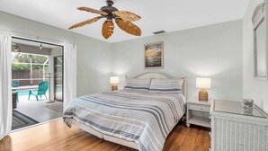 A bedroom with a bed, a ceiling fan, a nightstand, a lamp, and a view of an outdoor patio with a chair and a table. - A Quaint and Serene Neighborhood in North Naples (Naples)
