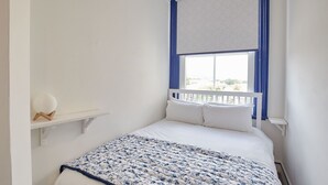Een slaapkamer met een bed, een raam met blauwe gordijnen, een witte muur en een klein plankje met een wit object.