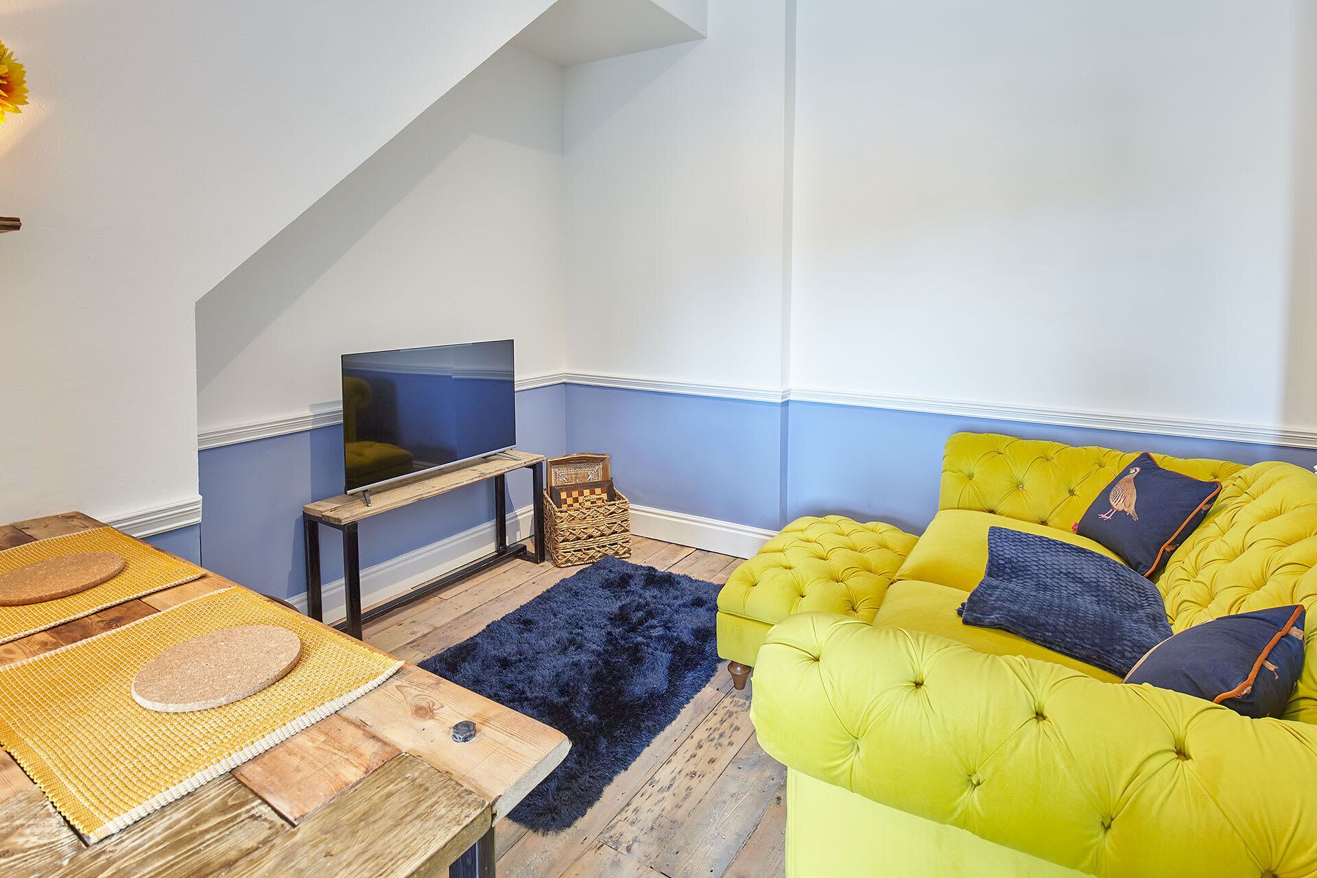 Een kamer met een geel sofa, een televisie op een houten standaard, een geweven mand en een donkerblauwe vloerkleed.