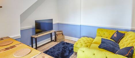 Een kamer met een geel sofa, een televisie op een houten standaard, een geweven mand en een donkerblauwe vloerkleed.