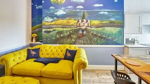 Een gele bank met uitzicht op een landschapspainting, een houten tafel en een keukenhoek.