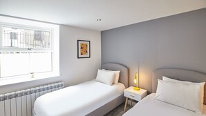 A hotel room with two beds, a window with a view of buildings, a radiator, and a bedside table with a lamp. - Host & Stay | The Royal Hideaway (Whitby)