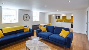 A modern living room with two blue sofas and a coffee table, a kitchen in the background, and a large window providing natural light. - Host & Stay | The Royal Hideaway (Whitby)