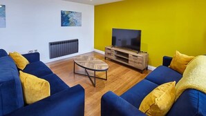 A modern living room with a blue sofa set, a wooden coffee table, and a flat-screen TV on a wooden stand. - Host & Stay | The Royal Hideaway (Whitby)