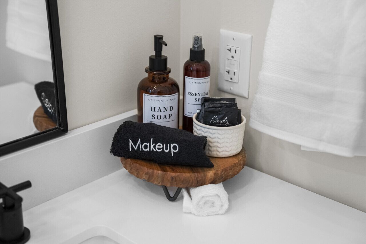 Un meuble-lavabo de salle de bain sur lequel repose un plateau en bois contenant une serviette noire étiquetée « Maquillage », un flacon de savon pour les mains et un récipient contenant des huiles essentielles.