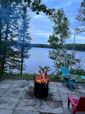A fire pit with logs, surrounded by chairs, overlooking a lake.