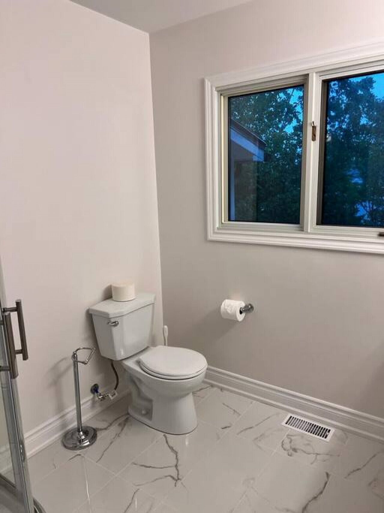 A bathroom with a toilet, a roll of toilet paper, a shower, and a window with a view of trees.