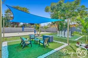 A backyard with artificial grass, a chess table, outdoor furniture, and a blue shade sail.
