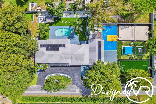 An aerial view of a resort with a swimming pool, tennis courts, and a landscaped area with trees and a fountain.