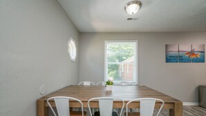 Un coin repas avec une table en bois et des chaises blanches, une fenêtre avec des stores et une œuvre d’art accrochée au mur.