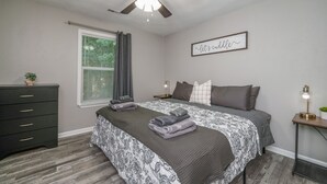 Une chambre à coucher comprenant un lit, une commode, un ventilateur de plafond, une fenêtre avec des rideaux, une table de chevet avec une lampe et un panneau décoratif mural.