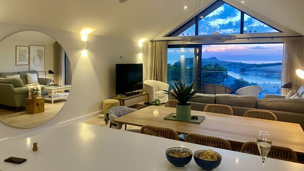 A modern living room with a large dining table, sofas, and a TV. There is a view of the ocean through large windows. - Porth Beach (Newquay)