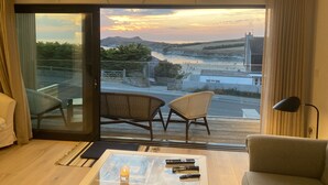 A modern room with a large glass door leading to a balcony, wicker chairs, a glass coffee table with remote controls, and a view of a coastal landscape at sunset. - Porth Beach (Newquay)