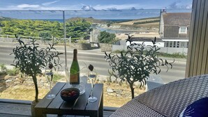A balcony with a table set for two, a bottle of wine, and a bowl of strawberries. The view includes a beach, a road, and a hill in the distance. - Porth Beach (Newquay)